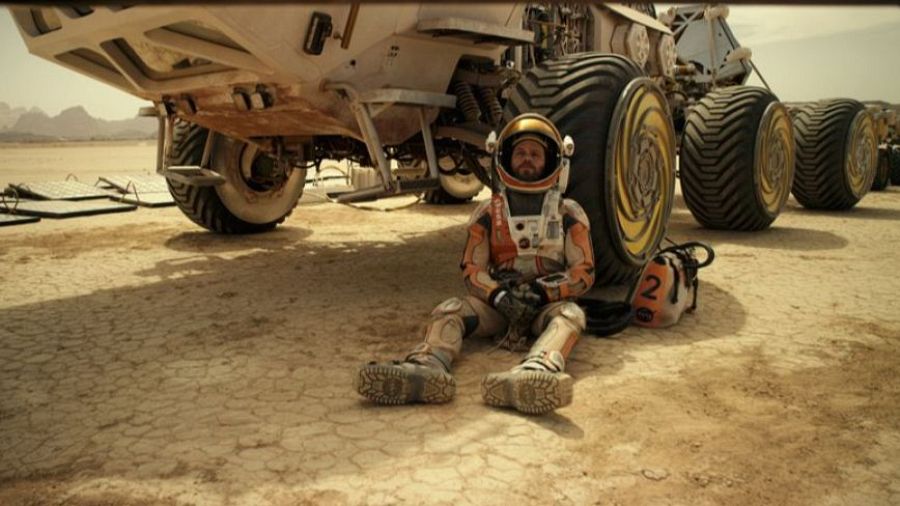 Un astronauta con un traje espacial naranja y blanco, y un casco dorado, se encuentra sentado en un terreno desértico y árido, con un vehículo de exploración blanco detrás. La escena sugiere un ambiente hostil y aislado.