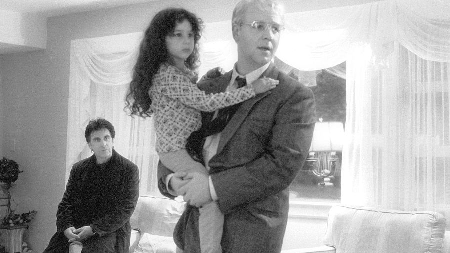 Una imagen en blanco y negro captura un momento íntimo: un hombre con traje sostiene a una niña, mientras otro hombre observa la escena en una habitación con cortinas y una lámpara.
