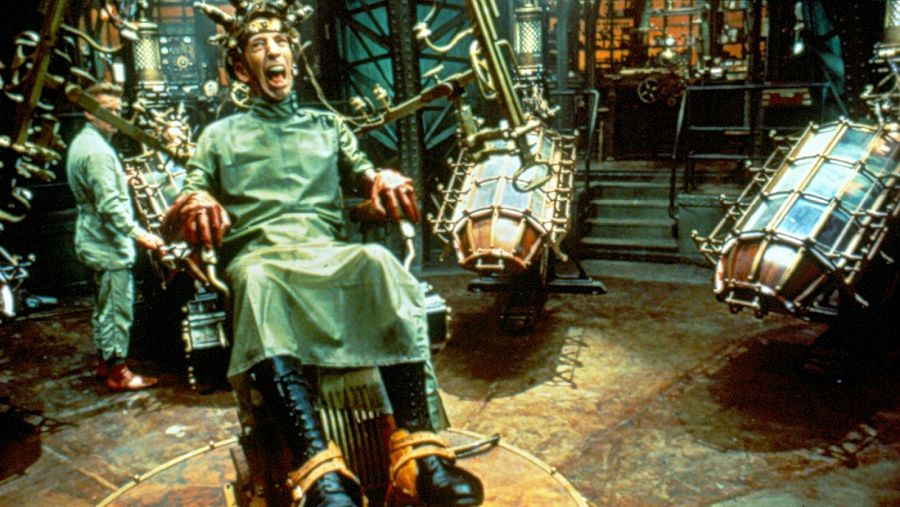 En un laboratorio con un diseño retrofuturista, un hombre con un dispositivo en la cabeza expresa terror mientras está sentado. Otro hombre, vestido con un uniforme, opera una máquina compleja en un entorno oscuro lleno de maquinaria.