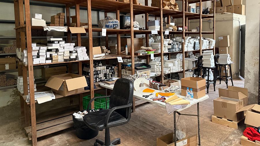 En el interior de un taller, una estantería de madera llena de cajas contrasta con una mesa de trabajo y una silla de oficina. Se observan materiales y objetos distribuidos por el espacio, sugiriendo actividad.