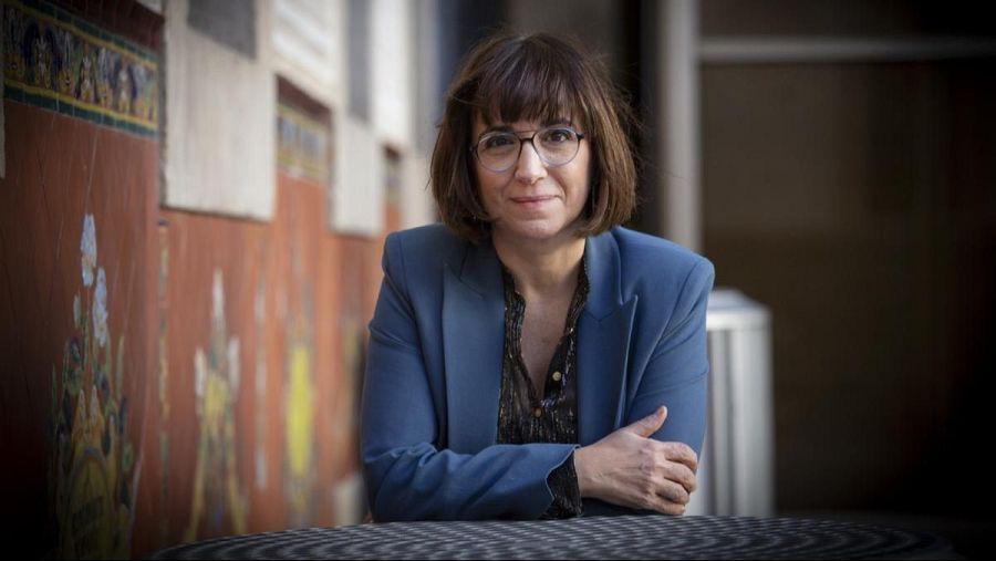 Judith Collel, con gafas y chaqueta azul, está sentada con las manos cruzadas. El fondo muestra una pared con paneles decorativos en tonos naranja y blanco.