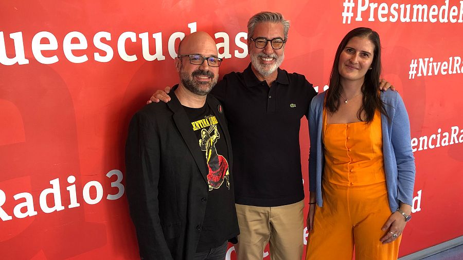 Tres personas posan en un estudio de radio: un hombre calvo con barba y gafas vestido de negro, otro con pelo canoso y gafas con un polo negro, y una mujer con mono naranja y chaqueta azul claro.
