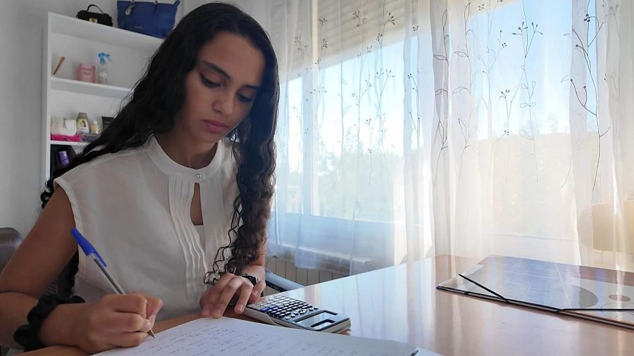 Una joven con cabello oscuro y blusa blanca escribe en un papel sobre una mesa, utilizando un bolígrafo azul. En la mesa también hay una calculadora. La habitación está iluminada por luz natural que entra por una ventana.
