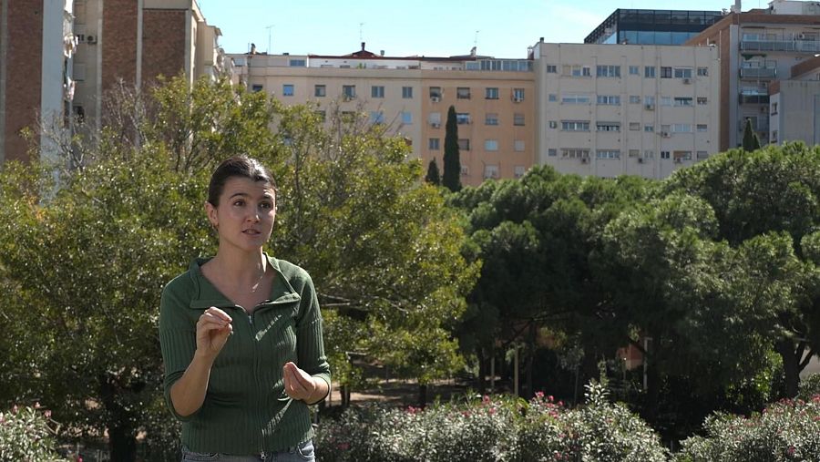 Carme Llogateres, con chaqueta verde y vaqueros, gesticula al aire libre, con edificios y vegetación al fondo. La imagen podría estar relacionada con una problemática social.