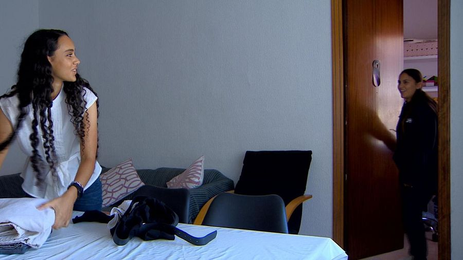 Dos chicas en un piso; una dobla ropa sobre una mesa con mantel blanco y chaqueta negra, mientras la otra entra por una puerta de madera.