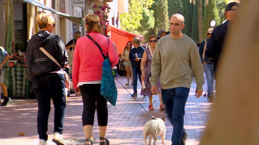 En una calle pavimentada con baldosas, un hombre con gafas y camiseta clara pasea a un perro blanco. Dos mujeres caminan detrás, una con chaqueta oscura y vaqueros, la otra con jersey rosa y pantalones oscuros, ambas con bolsos, mientras otras personas se dirigen en la misma dirección.