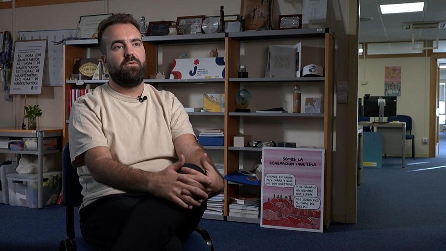 En una oficina, un hombre con barba y camiseta crema está sentado frente a una estantería con libros y objetos, donde se exhiben carteles con mensajes sobre la situación económica y la necesidad de manifestarse.