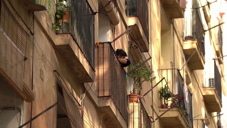Un edificio de apartamentos antiguo, con balcones y fachada deteriorada, es el escenario donde una persona se asoma a uno de ellos, apoyada en la barandilla, mientras la luz del sol ilumina parte de la construcción.