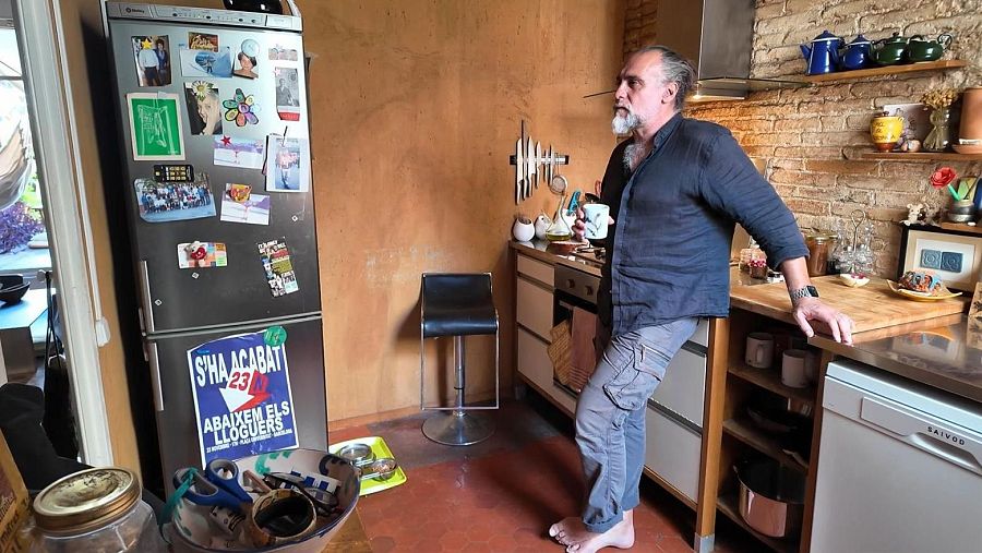 Un hombre de mediana edad, con barba canosa y camisa oscura, sostiene una taza en una cocina con paredes ocre y un refrigerador con imanes. Se observa una encimera con utensilios y una estantería con objetos decorativos.
