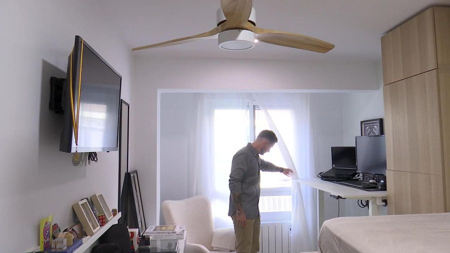 Un hombre joven, cerca de una ventana, ajusta una cortina en una habitación moderna. El espacio incluye un televisor, una estantería, un escritorio con ordenadores, una cama y un armario alto. Un ventilador de techo proporciona iluminación.