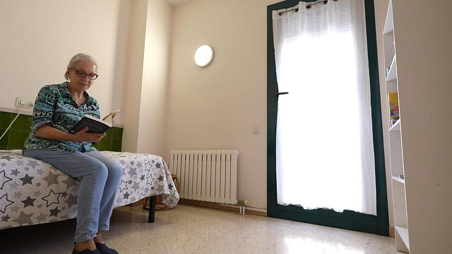 Una mujer mayor, Marisa, lee un libro sentada en una cama. La habitación, de aspecto sencillo, cuenta con una cama con colcha estampada, azulejos verdes y una ventana con cortinas.