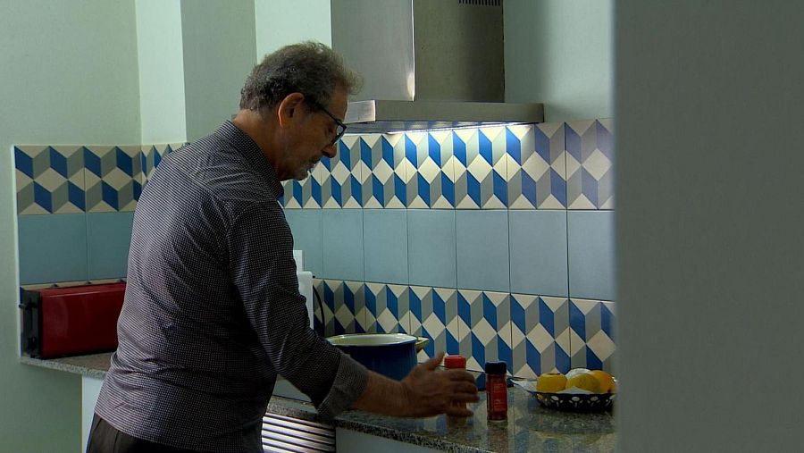 Un hombre mayor, Ramón Cuina, con gafas y camisa oscura, cocina en una cocina. En la encimera, se observan una olla azul, botes de especias y limones, con azulejos geométricos al fondo.