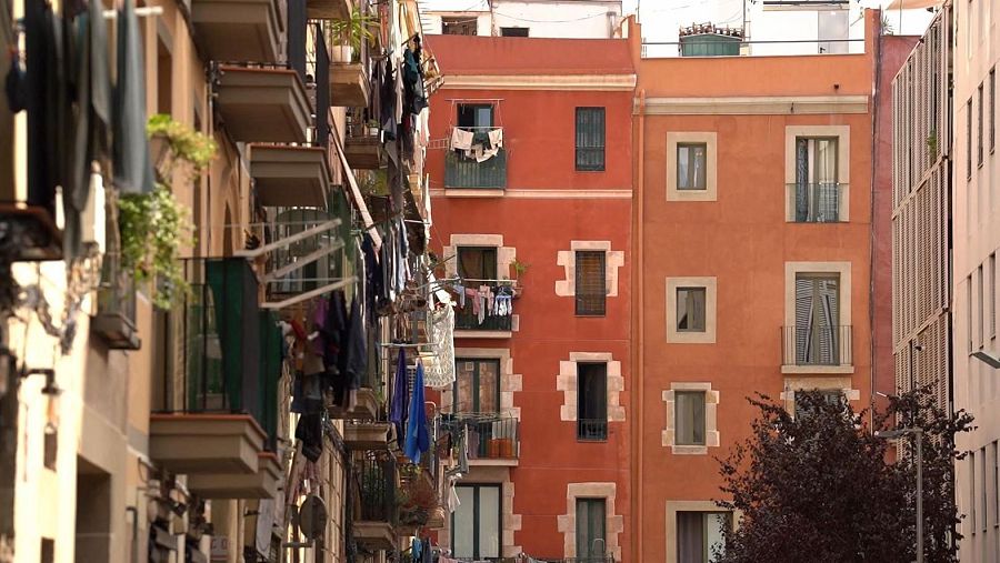 Una calle con edificios coloridos y balcones con ropa tendida. El texto "EDIFICIS ROBA ESTESA Informe Semanal" destaca la problemática de la vivienda, mencionando el alquiler por habitaciones y la disminución de la calidad de vida.