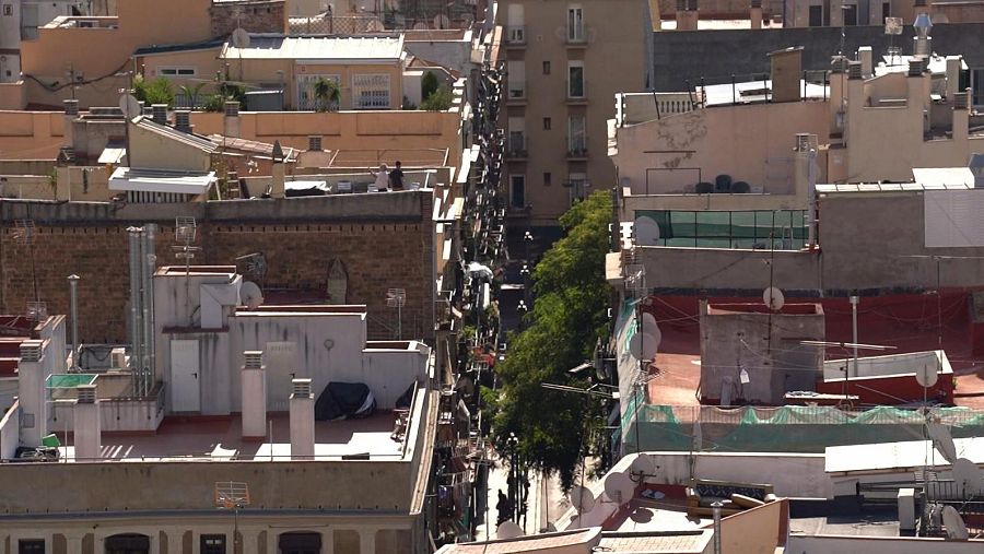 Vista aérea de una calle urbana con edificios de diferentes alturas y estilos, con azoteas equipadas y personas caminando. Se distingue el texto "TEULADES" y "Informe Semanal".