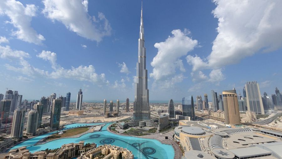 El Burj Khalifa, un rascacielos imponente, se alza sobre el paisaje urbano, rodeado de otros edificios y fuentes, con un cielo azul y nubes blancas como telón de fondo.