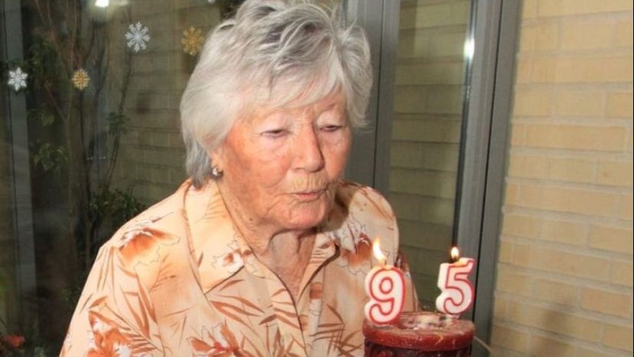 Una mujer mayor, con el pelo canoso y una camisa estampada, sopla las velas de una tarta de cumpleaños con el número 95. Se encuentra en una habitación con una ventana decorada.