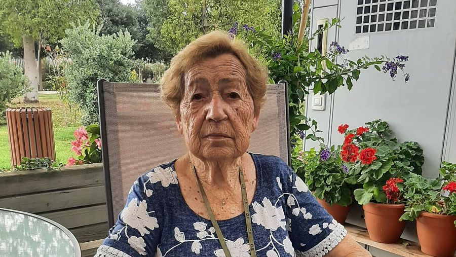 Una mujer mayor, identificada como Rosa, está sentada en una silla al aire libre, vestida con una blusa estampada y un cordón. El fondo muestra vegetación, macetas con flores y una pared con rejillas.