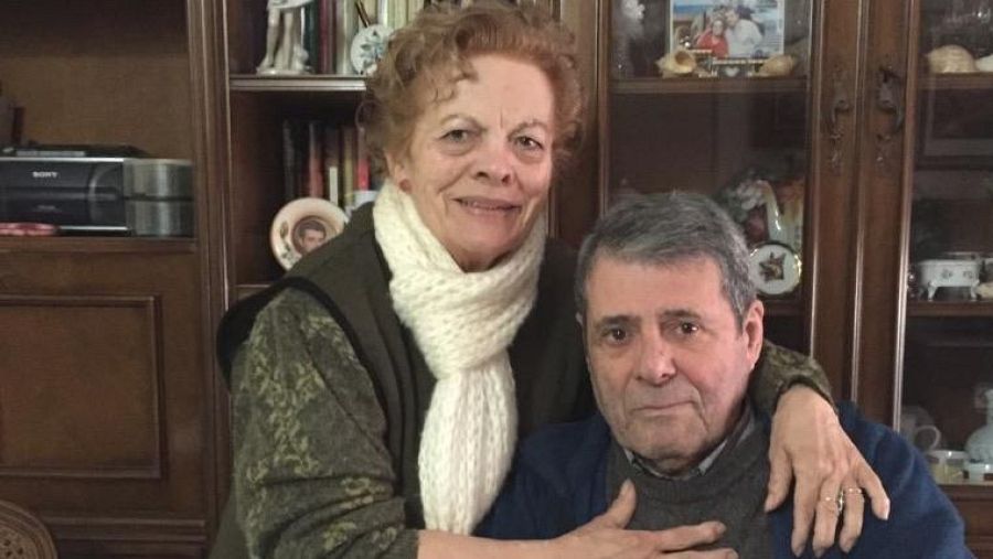 En el interior de una vivienda, un hombre con un jersey azul y una camisa gris está sentado, mientras una mujer con una blusa estampada y una bufanda blanca lo abraza. Se observa un mueble de madera con libros y adornos.