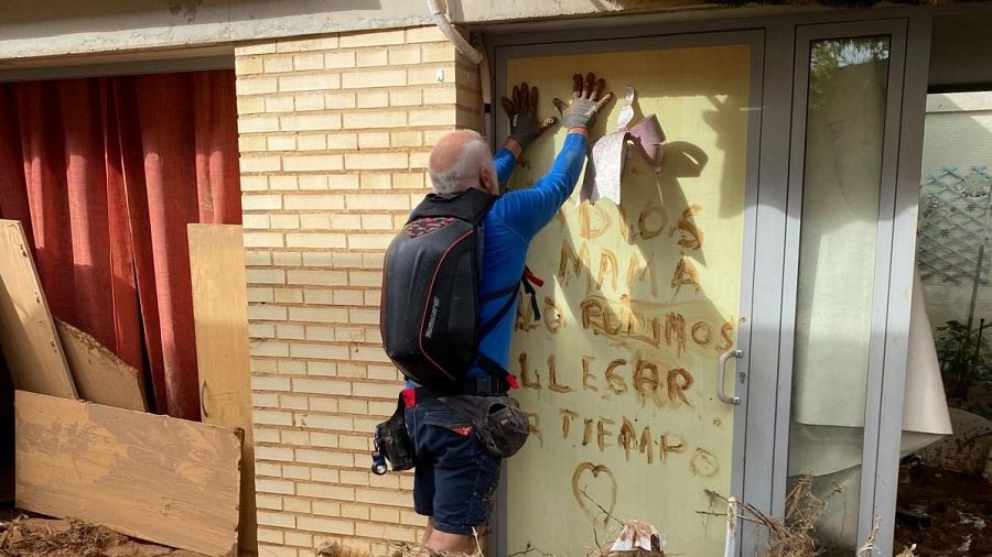 Un hombre con camiseta azul y mochila negra escribe un mensaje de despedida en una puerta de cristal embarrada, posiblemente tras un desastre. El mensaje, escrito con barro, incluye un corazón.