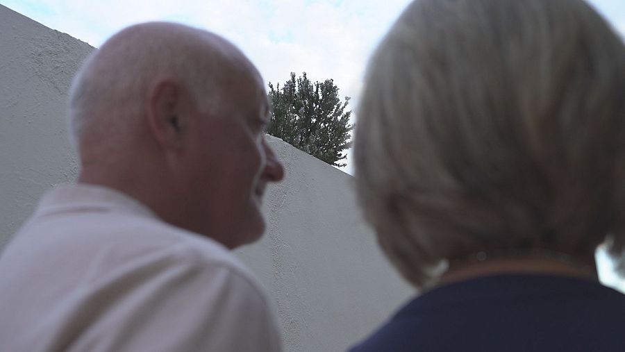 Una pareja camina frente a una pared blanca; el hombre, de perfil, con camisa clara y cabeza calva, y la mujer, de espaldas, con blusa oscura y pelo canoso. Un arbusto se encuentra detrás de la pared.