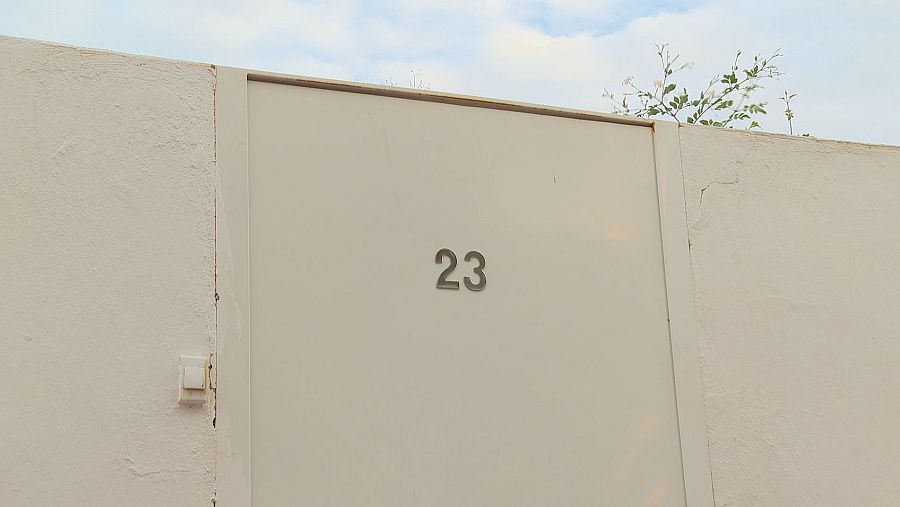Una puerta blanca, con el número 23 en relieve, se alza solitaria. Enmarcada por una pared blanca con textura, la puerta se encuentra bajo un cielo visible y vegetación.