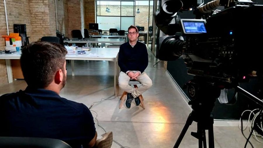 En un espacio con paredes de ladrillo visto y suelo de cemento pulido, un periodista con gafas y jersey azul es grabado por un cámara con equipo profesional.