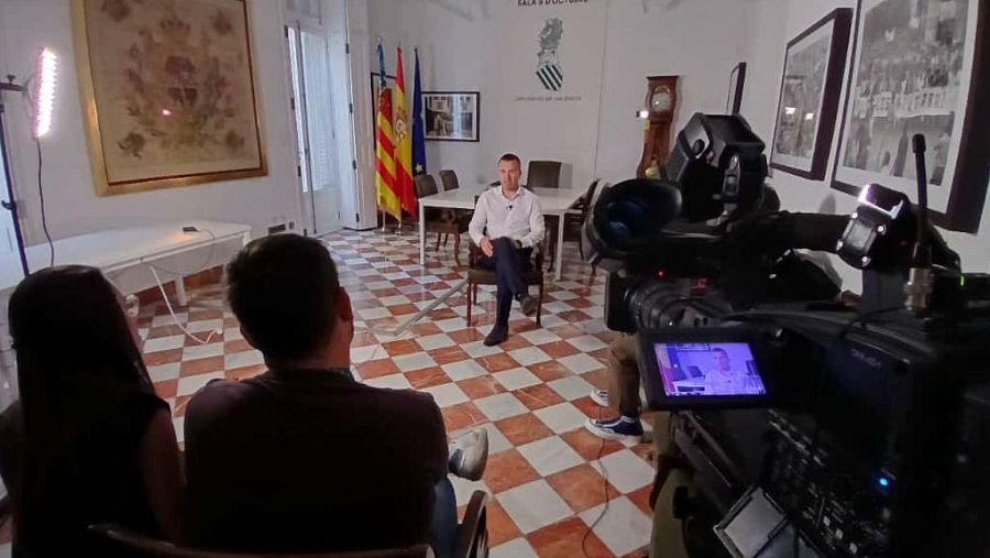 En un plató de rodaje, un hombre sentado con camisa blanca y pantalones oscuros es el foco de atención. Tras él, se observan banderas y una mesa con sillas, mientras una cámara de vídeo profesional graba la escena.