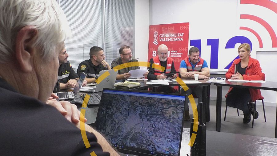Una reunión en una sala muestra a varias personas alrededor de una mesa, incluyendo a individuos con uniformes y una mujer con chaqueta roja. Un ordenador portátil en primer plano exhibe un mapa detallado.