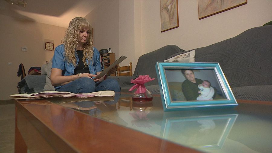 Una mujer rubia, vestida con camisa azul y vaqueros, está sentada en el suelo de una sala, mirando una fotografía. A su alrededor hay papeles y, frente a ella, una mesa con un marco de fotos y un jarrón.