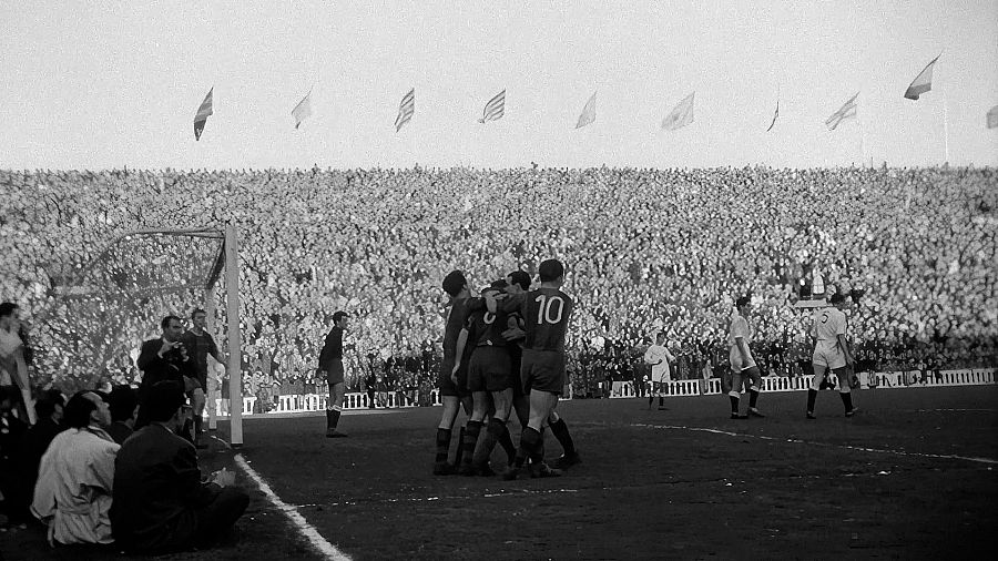 En primer pla, un grup de jugadors celebra un gol en un estadi ple de públic. Un jugador porta el número 10 a la samarreta. Al fons, les grades estan plenes d'espectadors.
