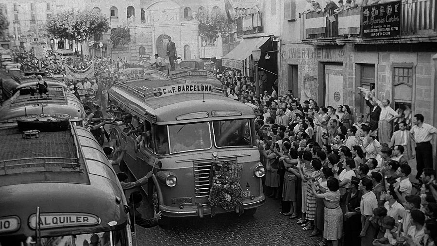 Una multitud celebra el pas d'autobusos decorats amb motius esportius. Els vehicles, encapçalats per un amb una corona de flors, avancen per un carrer ple de gent, en una escena festiva.