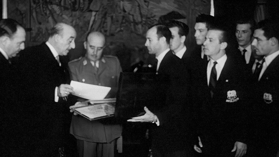 Francisco Franco, en uniforme militar, rep una delegació del FC Barcelona al Palau del Pardo. Els jugadors, amb vestits foscos i corbates, li presenten documents, possiblement relacionats amb el club.