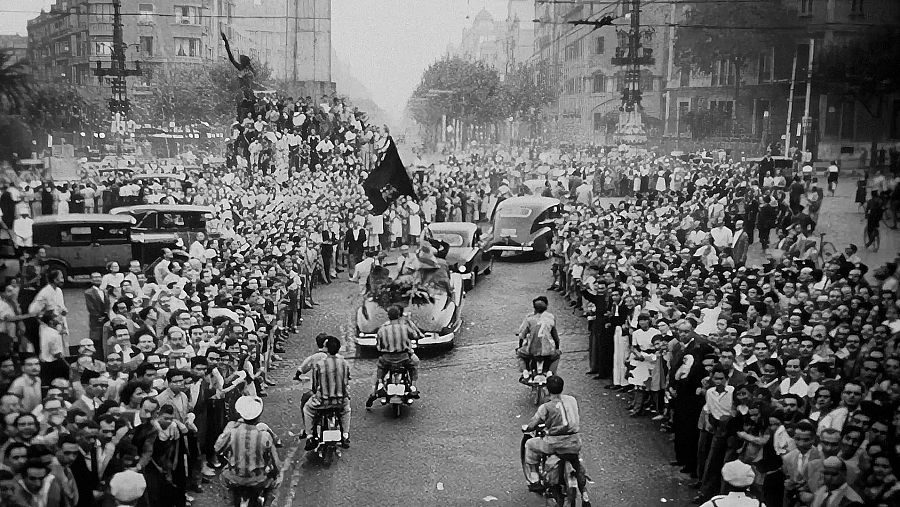 Una escena en blanc i negre mostra una multitud a banda i banda d'un carrer, amb cotxes i motocicletes clàssiques desfilant lentament. La gent, vestida amb roba d'època, sembla estar celebrant o participant en un esdeveniment públic.