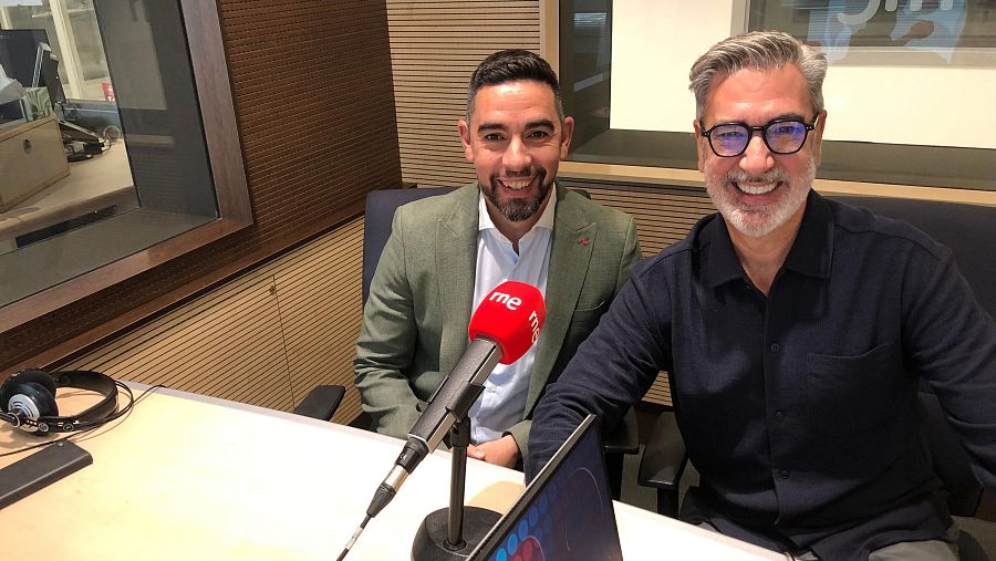 Dos hombres sonríen en un estudio de radio, sentados frente a micrófonos. Uno viste camisa clara y chaqueta verde, el otro camisa oscura y gafas. En la mesa hay auriculares y una tablet.