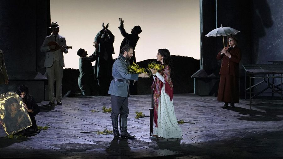 Una mujer vestida de blanco con un chal rojo recibe un ramo de flores de un hombre con uniforme militar, mientras un músico toca la mandolina y dos figuras observan la escena teatral.