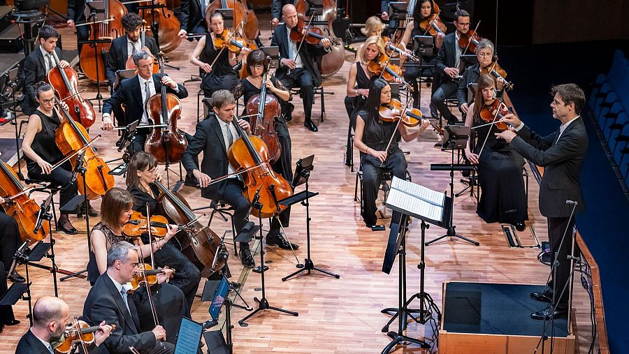 Una orquesta sinfónica, compuesta por músicos con instrumentos de cuerda, actúa en un escenario bajo la batuta de un director. El público, ubicado en una sala de conciertos, disfruta de la interpretación musical.