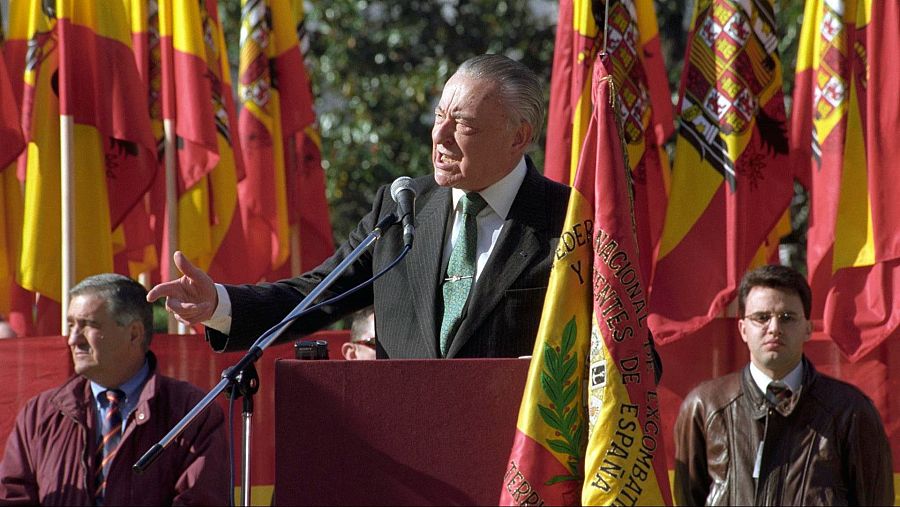 Blas Piñar, con traje oscuro y corbata verde, habla ante un micrófono en un acto público, con banderas de España de fondo. A su lado, dos hombres observan la escena.