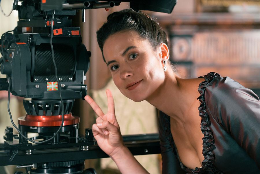 En el set de rodaje, una mujer sonriente con un vestido oscuro y el pelo recogido celebra con el signo de la victoria, junto a una cámara de cine profesional con una pegatina.