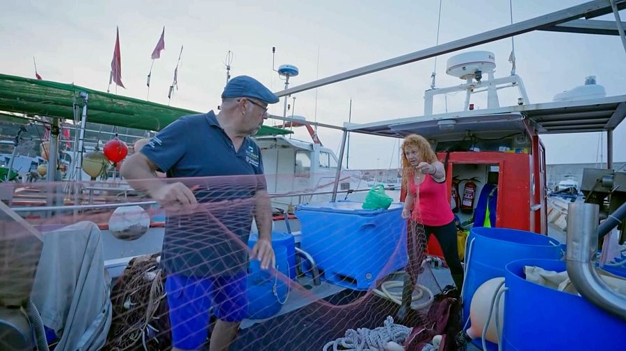 En una escena de pesca, un home amb gorra blava i una dona amb samarreta vermella col·laboren en la manipulació d'una xarxa a la coberta d'un vaixell, amb contenidors blaus i altres equips a la vista.