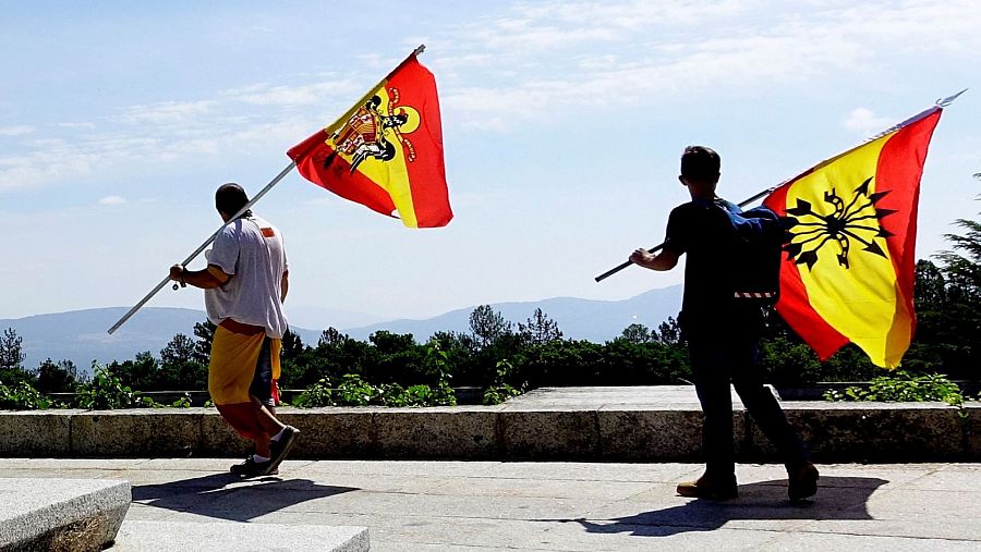 En un paisaje montañoso, dos hombres sostienen banderas con simbología preconstitucional. Uno lleva una bandera con el escudo de España y el otro, una con el yugo y las flechas.
