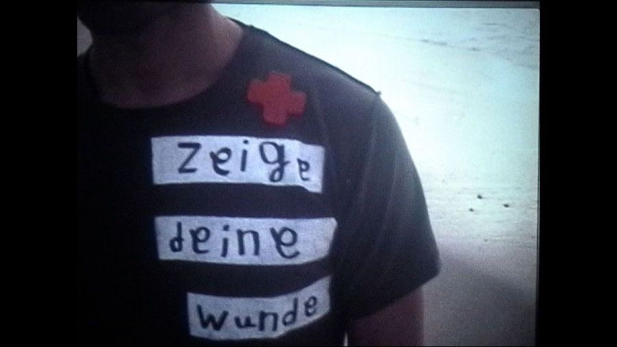 Una persona viste una camiseta negra con la frase en alemán "Zeige deine Wunde" escrita en blanco, y una cruz roja en el hombro, en un entorno costero.