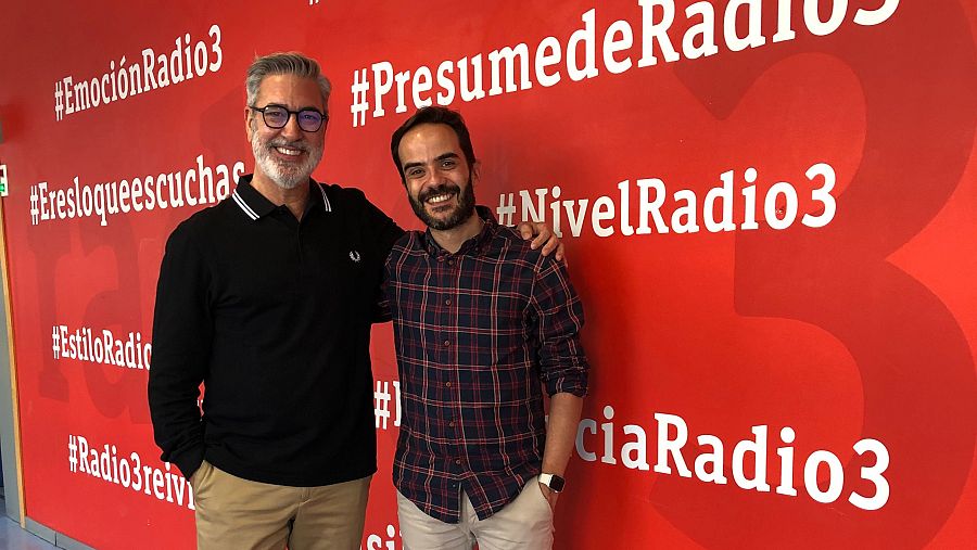 Dos hombres posan juntos en un estudio de radio. Uno de ellos lleva gafas y un polo negro, mientras que el otro viste una camisa de cuadros. El fondo es rojo con hashtags relacionados con una emisora.