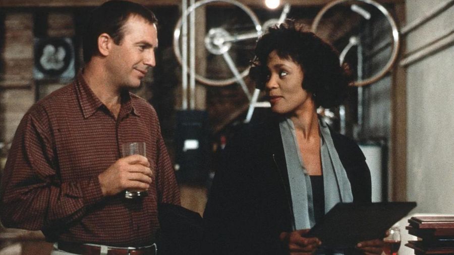 Kevin Costner y Whitney Houston comparten un momento en un espacio interior. Costner sostiene un vaso, mientras Houston tiene un documento en sus manos. El entorno sugiere un almacén o garaje.