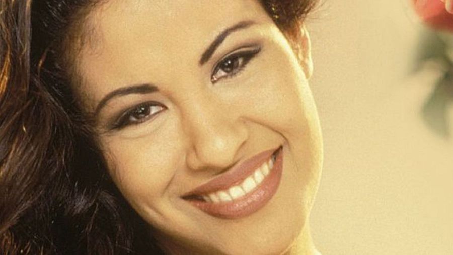 Primer plano del rostro de una mujer sonriente con cabello oscuro, ojos marrones delineados y piel clara. Se aprecia una flor roja desenfocada en el fondo.