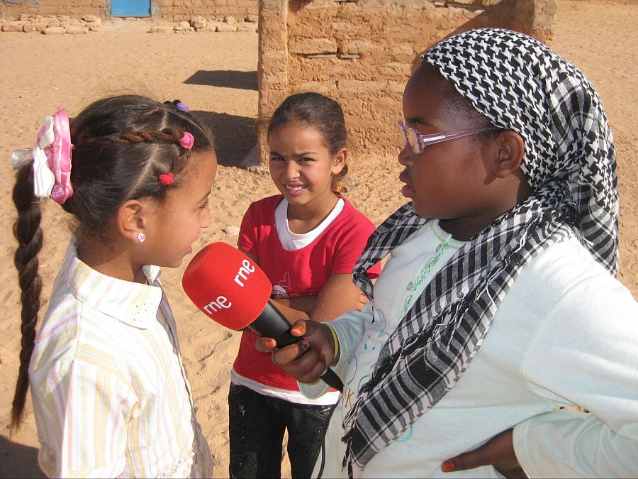 En un campamento desértico, dos niñas saharauis participan en una entrevista. Una de ellas, con una trenza y camisa a rayas, es entrevistada mientras otra sostiene un micrófono con el logo de "rne". Una tercera niña sonríe en segundo plano.