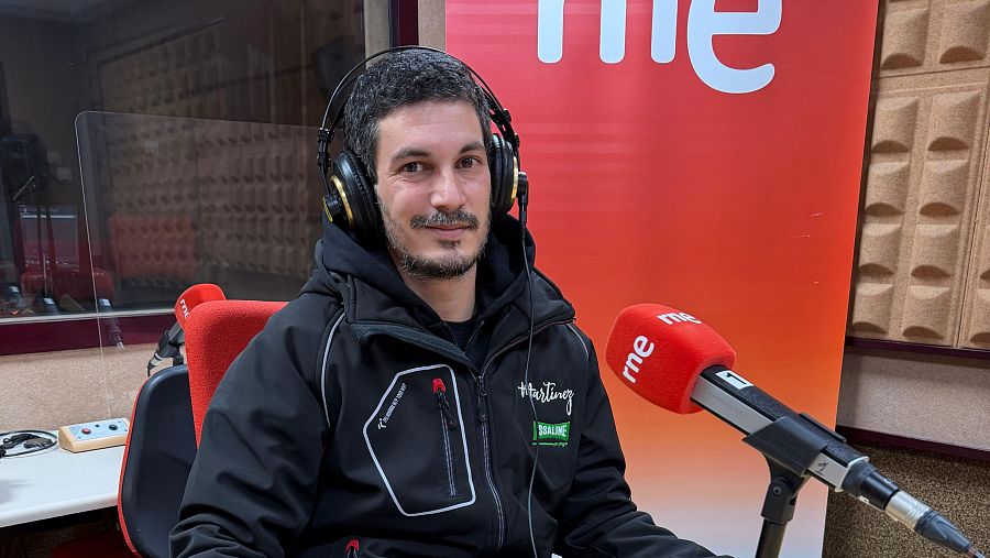Joel Martínez, en los estudios de radio, posa mirando a la cámara con auriculares y una chaqueta con logos. Un micrófono rojo y un banner con el logo de la emisora completan la escena.