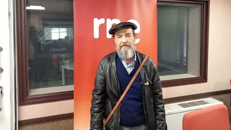 Un hombre con barba y gorra, posa frente a un panel con un logo. Viste una chaqueta de cuero, un jersey y una camisa a cuadros, con una bandolera. Se observa una ventana con una persona al fondo.