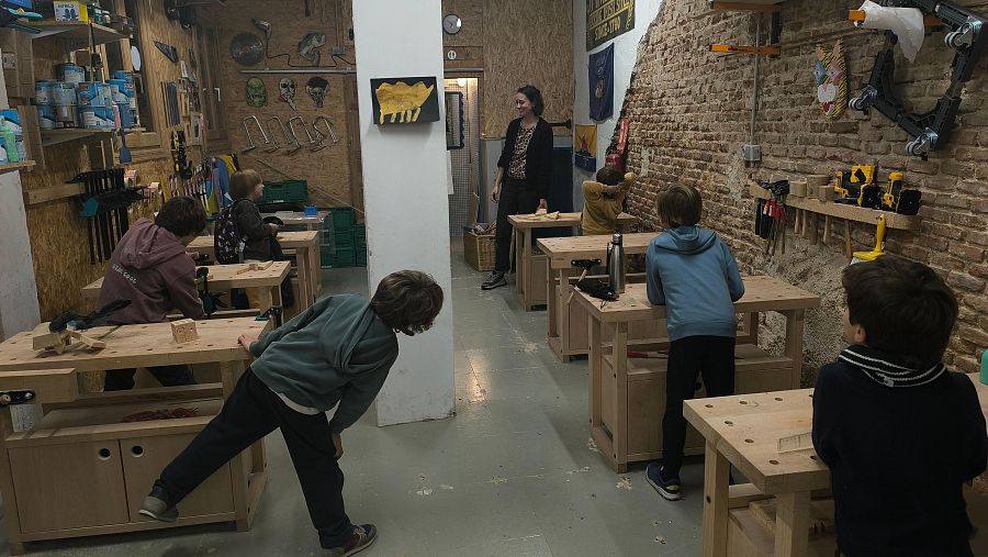 Ana Rodríguez enseña ebanistería a un grupo de niños en un taller. Los niños, concentrados, manipulan herramientas y trabajan la madera en mesas, rodeados de herramientas y objetos de madera.