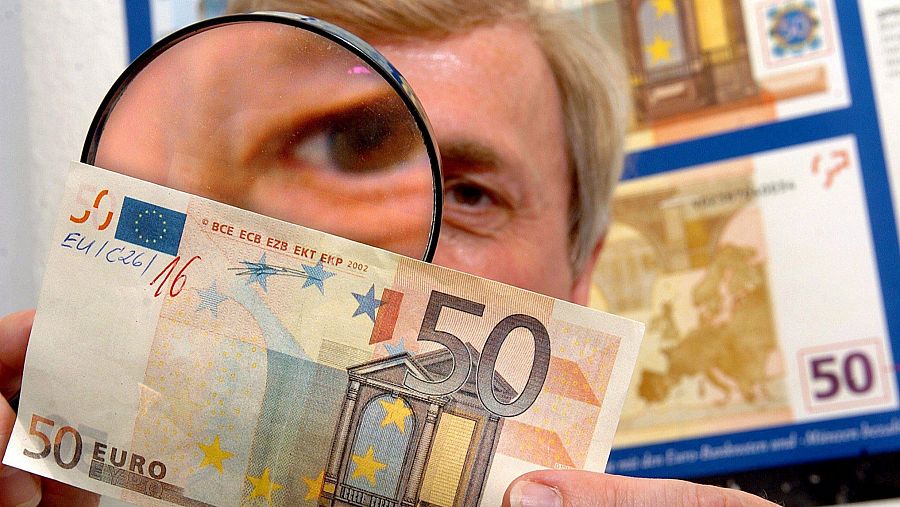 Una persona utiliza una lupa para examinar un billete de 50 euros, enfocando una sección donde se aprecia el número "50" y detalles del diseño.