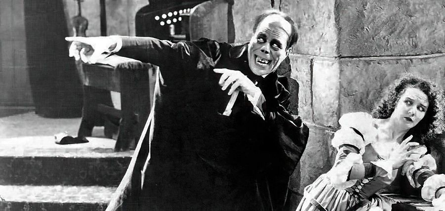 En una imagen en blanco y negro, un hombre con maquillaje grotesco y vestimenta oscura, posiblemente un personaje de ficción, extiende un brazo con gesto amenazante. A su lado, una mujer con el rostro aterrorizado observa la escena. El fondo sugiere un entorno oscuro y antiguo.